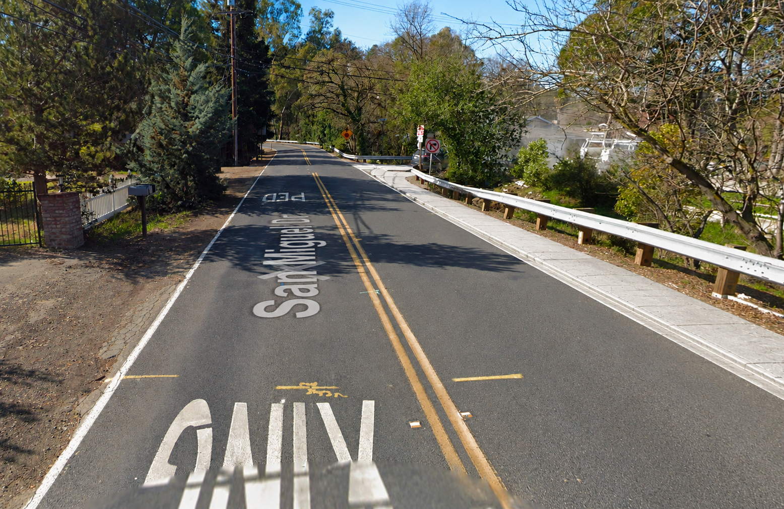 A view down San Miguel Drive showing one narrow shoulder with a guardrail and no continuous sidewalk on either side